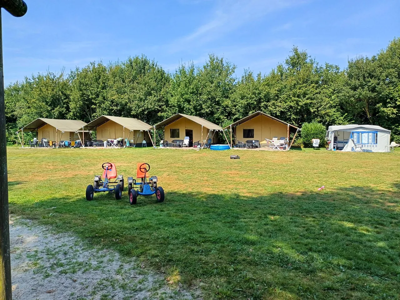 Vier safaritenten op een grasveld voor een bosrand, met twee skelters op het gras