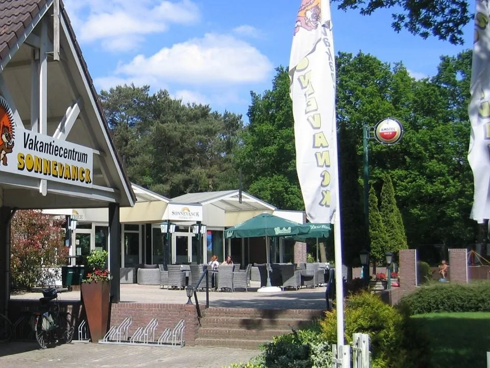 Ingang en terras van Vakantiecentrum Sonnevanck met vlaggen en tuinmeubilair onder bomen